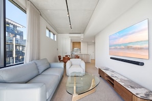 Uma sala moderna com uma mesa de centro de vidro, um sofá, um espaço de jantar com cadeiras e uma cozinha ao fundo.