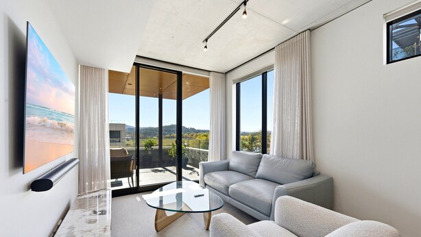 A modern living room with a glass coffee table, grey sofa, and a large window offering a view of the outdoors. - Tranquil 2-bed With Water View (Canberra)
