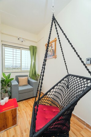 Een kamer met een zwarte hangstoel, een grijze bank met een kussen, een houten tafel met een rode tafelkleed, en een raam met groene gordijnen.