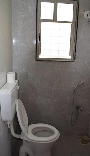 A bathroom with a toilet, a roll of toilet paper, a shower, and a window with a grid pattern. - Vibestayz Omkar Hinjawadi (Pune)