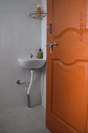 A bathroom with a white sink, a wooden door, and a glass shelf with toiletries. - Vibestayz Omkar Hinjawadi (Pune)