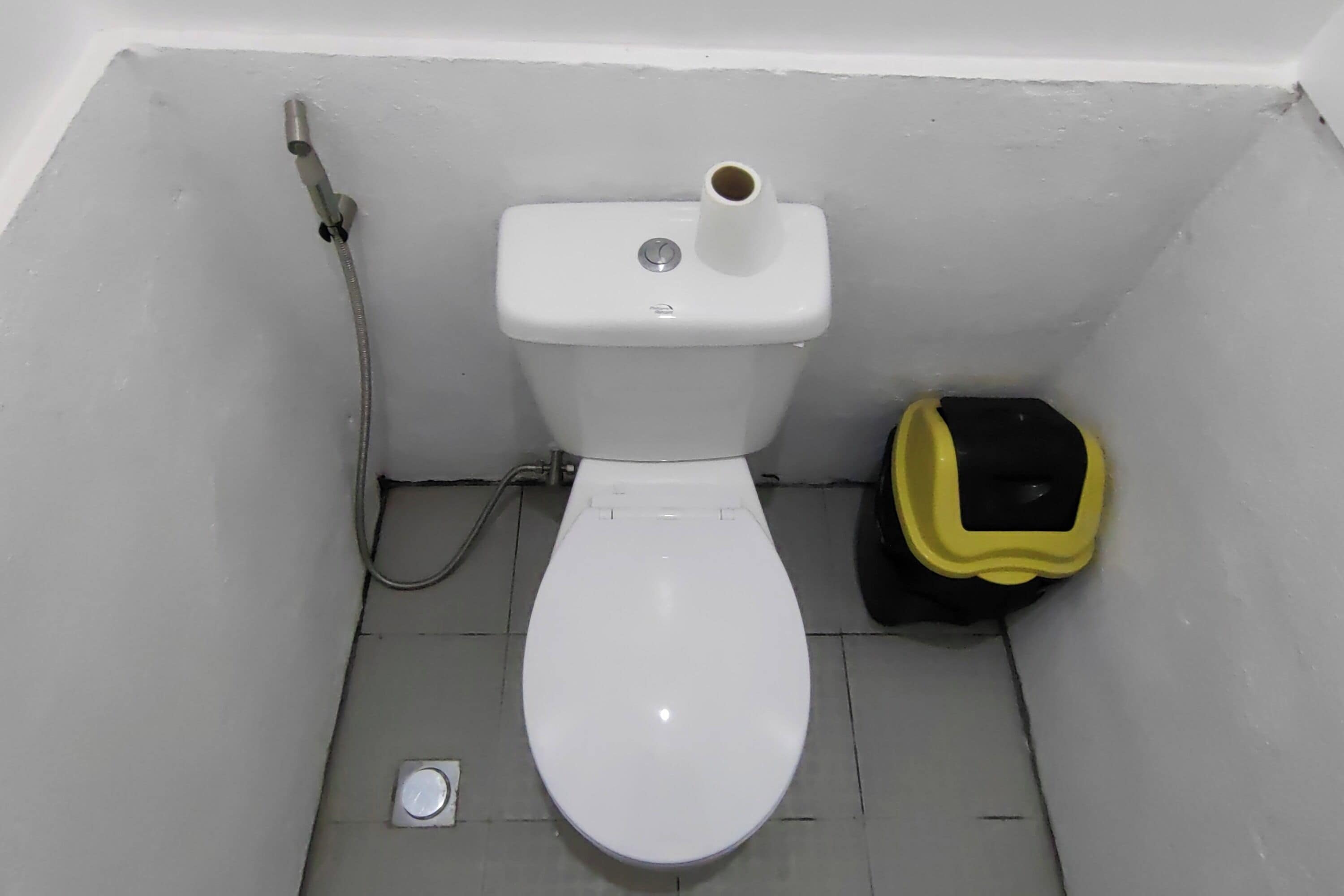 Un inodoro blanco con un bidé, un cesto de basura pequeño y un desagüe en el suelo.