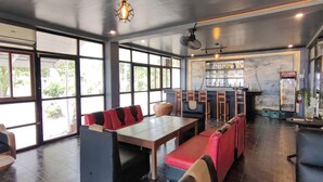 A restaurant with a bar area, seating with red and black cushions, and a large window providing a view of the outdoors. - Puerto Paraiso Hostel (Puerto Princesa)