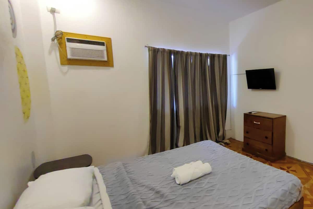 Un dormitorio con cama, mesita de noche, cómoda, televisor montado en la pared y ventana con cortinas.