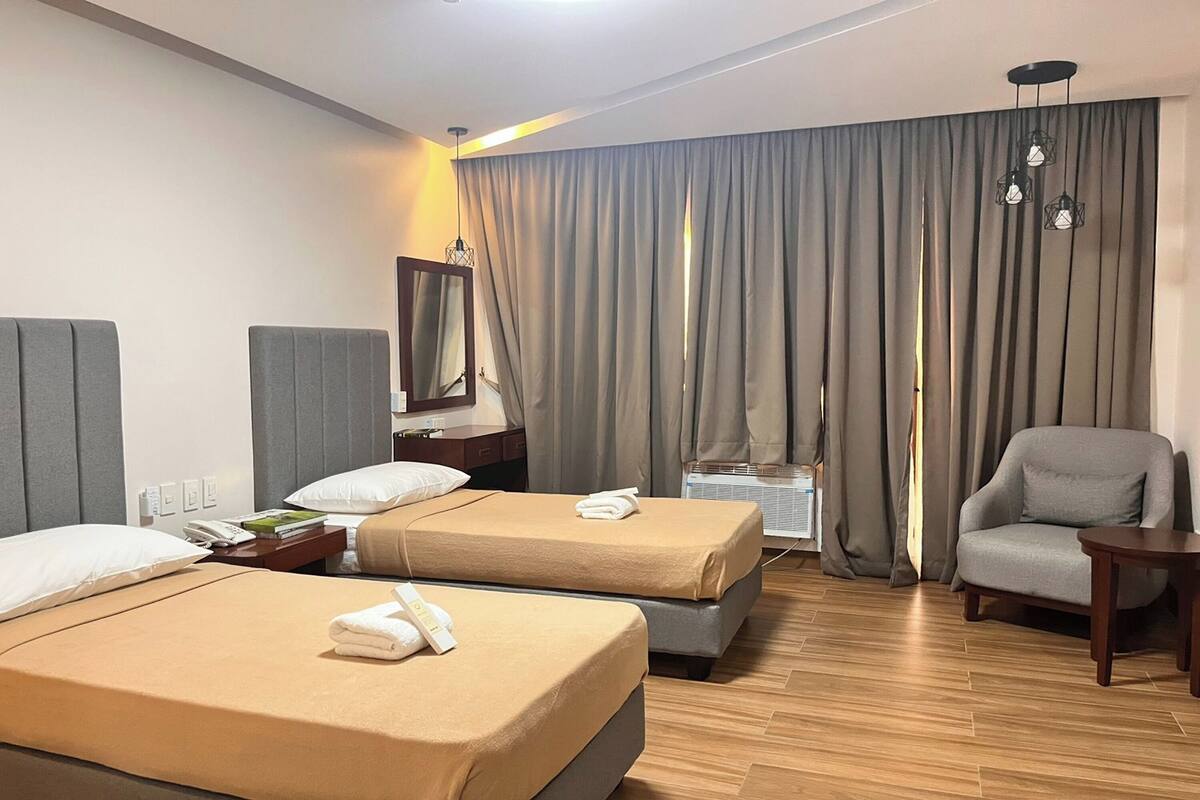 Hotellihuone, jossa on kaksi sänkyä, tuoli, pieni pöytä, peili ja ikkuna, jossa on verhot.