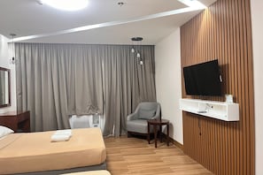 A hotel room with a bed, a chair, a TV mounted on a wooden panel, and a window with curtains.