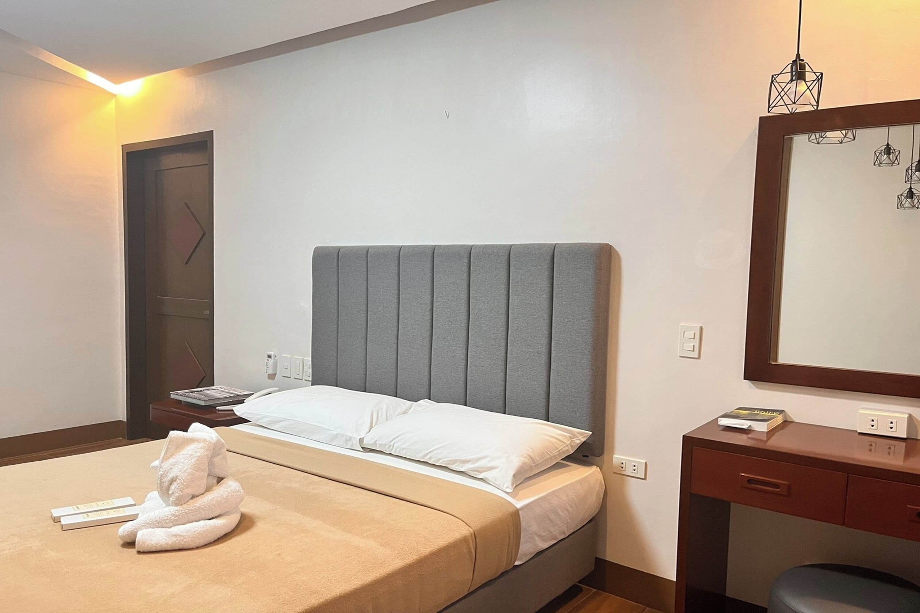 Una cama bien hecha con cabecera gris, una mesita de noche con un libro, un escritorio pequeño con una silla y un espejo en la pared.