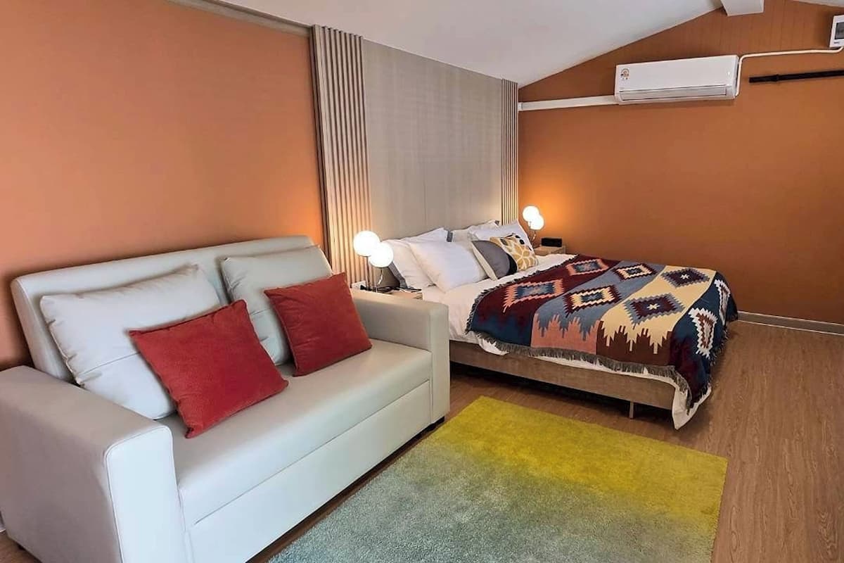 A hotel room with a bed, a sofa, a bedside table, an air conditioner, and a window with curtains.