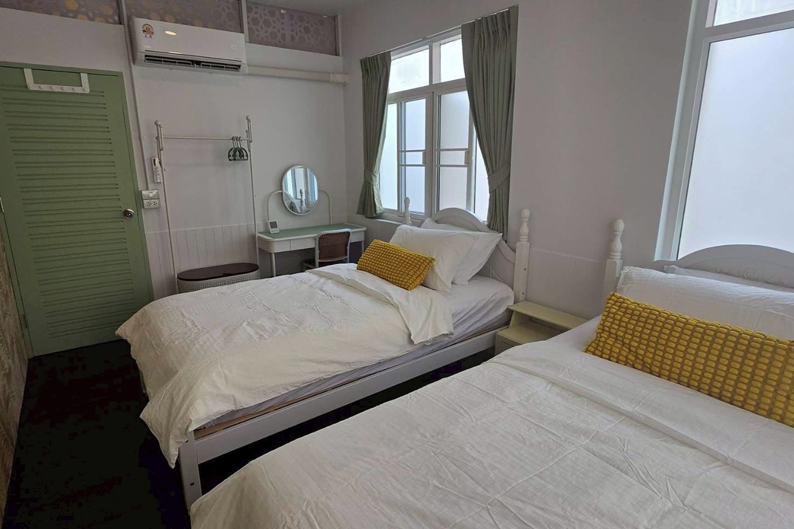 A room with two beds, a desk with a mirror, a window with curtains, and an air conditioning unit.