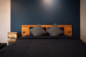 Un dormitorio con pared azul oscuro, cabecera de madera, cama con ropa de cama oscura y una lámpara de noche.