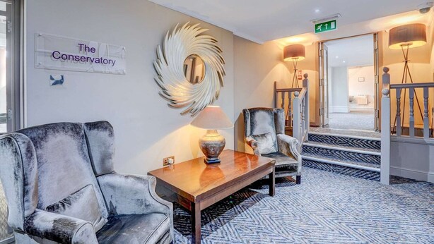 Un vestíbulo de hotel con un cartel que dice 'The Conservatory', un espejo decorativo grande, una mesa de centro de madera, un sofá, una silla y una escalera con barandilla.