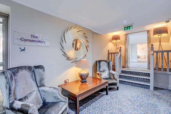 En hotellobby med en skylt som lyder ”Orangeriet”, en stor dekorativ spegel, ett träbord, en soffa, en stol och en trappa med ett räcke.