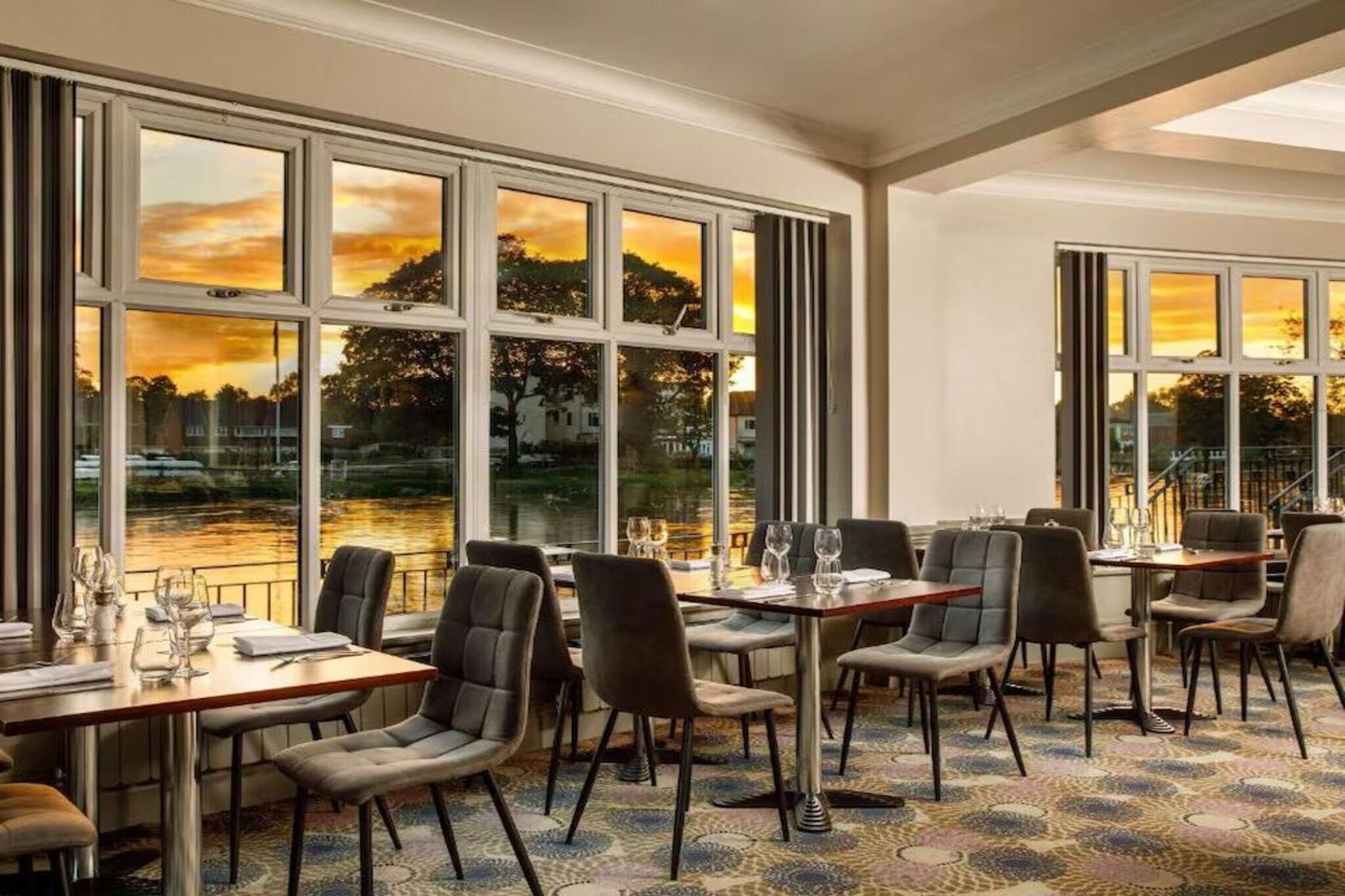 A dining area with tables and chairs, large windows with a view of a sunset over a body of water.