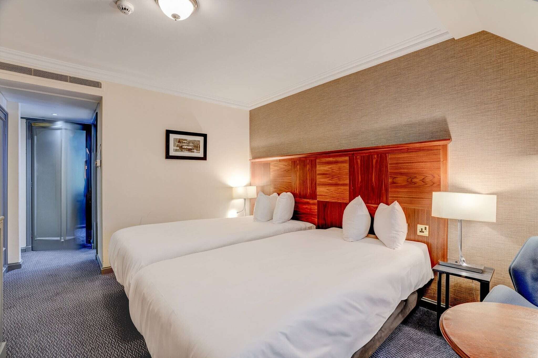 A hotel room with two beds, a wooden headboard, a framed picture on the wall, and a desk with a lamp.