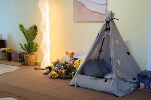 Un espacio acogedor con zona de descanso que incluye una estructura tipo tienda, una lámpara, peluches y una planta en maceta.