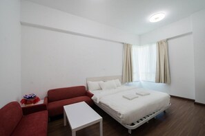 Una habitación de hotel con una cama, un sofá rojo, una mesita blanca y una ventana con cortinas.