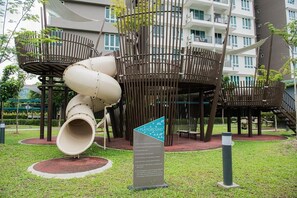 A playground with a slide and climbing structures, surrounded by a grassy area and a building in the background.