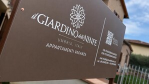 En skylt för ”Giardino delle Ninfe” med en QR-kod, kontaktinformation och information om ”Appartamenti Vacanza” i Umbrien, Italien.