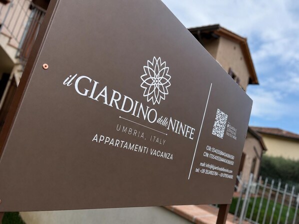 A sign for 'Giardino delle Ninfe' with a QR code, contact information, and mentions 'Appartamenti Vacanza' in Umbria, Italy.