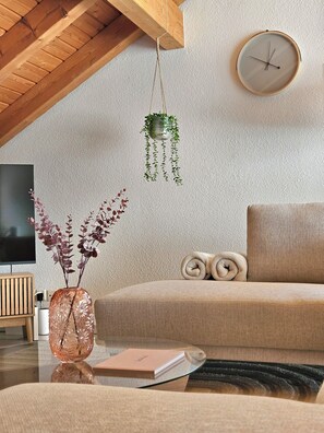 A living room with a sofa, a round wall clock, a hanging plant, and a vase with dried flowers. - 4Seasons | Morschach | Family Apartment | Mountain View (Morschach)