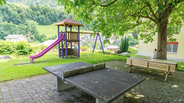 Une aire de jeux avec un toboggan et des balançoires, un banc et une zone dallée.