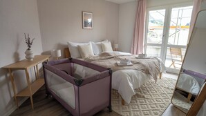 Une chambre à coucher comprenant un lit, un berceau, une table de chevet, une lampe, un tapis et une fenêtre donnant sur un beau paysage.