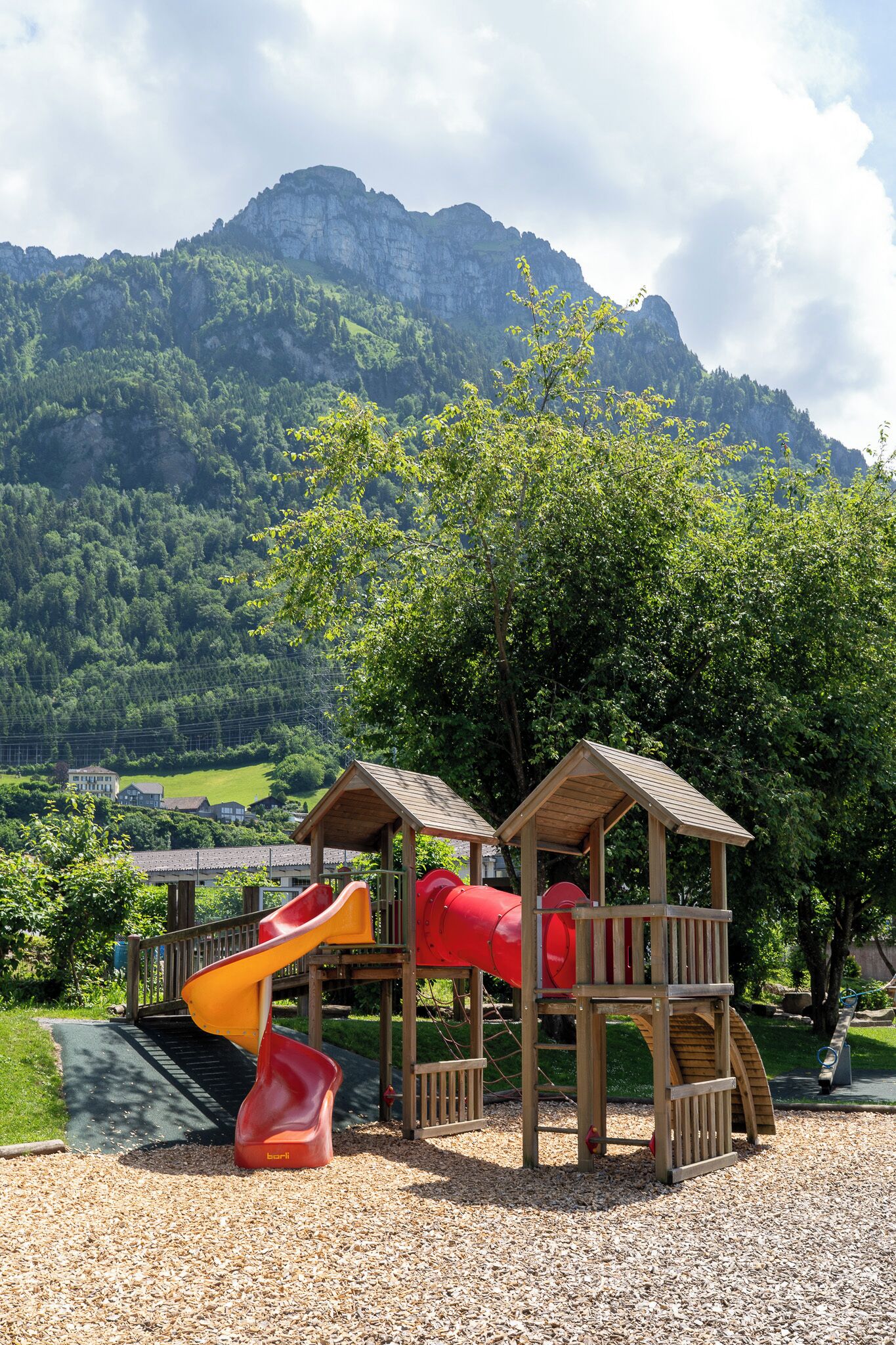 Ein Spielplatz mit Rutsche und Klettergerüsten, vor einer Berg- und Baumlandschaft.