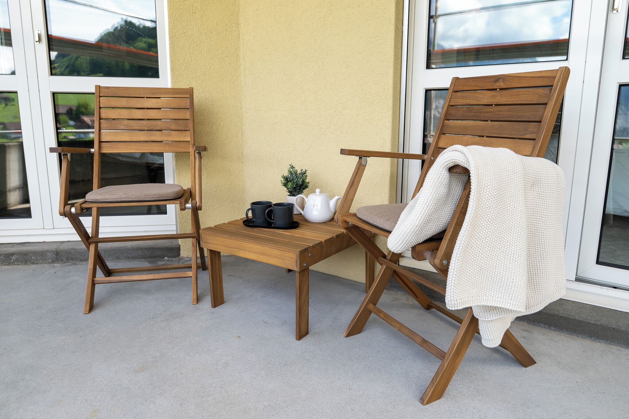 Deux chaises en bois avec des coussins, une petite table, une théière blanche et deux tasses noires sur un balcon dont le sol est en béton et le mur est jaune.
