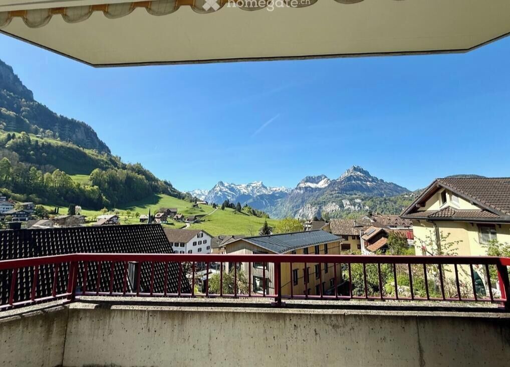 Un balcon avec une balustrade rouge donnant sur un paysage montagneux, avec des maisons et un ciel dégagé.