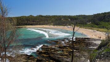 A coastal beach with clear turquoise waters, rocky shoreline, and a forested background.