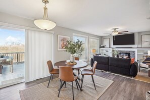 A modern living room with a fireplace, a dining area with a round table and chairs, and a balcony with outdoor furniture.