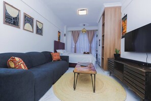 Ein gemütlicher Wohnraum mit Sofa, einem kleinen Tisch, einem Bett, einem Fernseher und Kunstwerken an den Wänden.