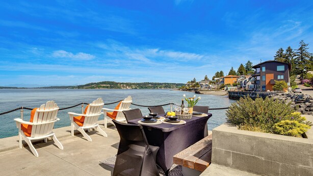 A waterfront dining area with chairs and a table, overlooking a body of water and buildings. - Waterfront Cabin w/ Stunning Sound Views (University Place)