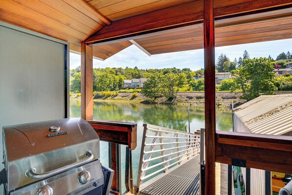 A view from a covered deck overlooking a river, with a grill and a railing.
