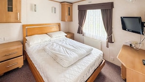 Ein ordentlich bezogenes Bett mit weißer Steppdecke, ein Holzkopfbrett, ein Nachttisch mit Lampe, ein Kleiderschrank, ein an der Wand befestigter Fernseher und ein Fenster mit Vorhängen.