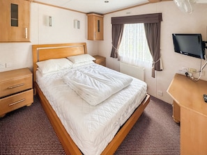 Ein ordentlich bezogenes Bett mit weißer Steppdecke, ein Holzkopfbrett, ein Nachttisch mit Lampe, ein Kleiderschrank, ein an der Wand befestigter Fernseher und ein Fenster mit Vorhängen.