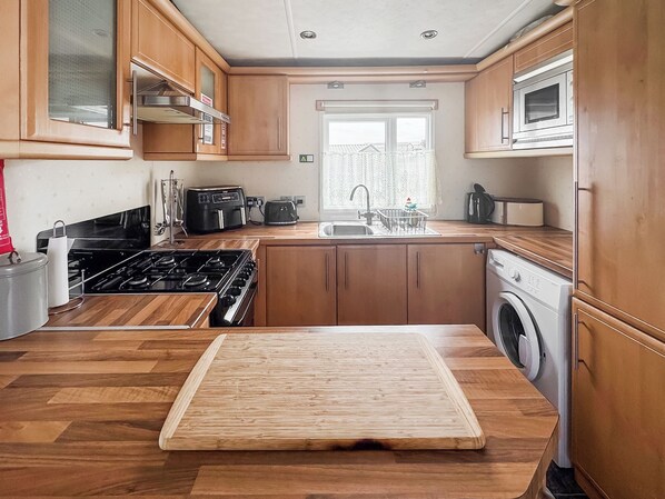 A kitchen with wooden cabinets, a gas stove, a microwave, a washing machine, and a window with a view of houses.