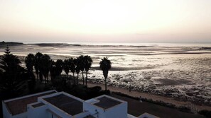 A coastal view with a beach, palm trees, and a building with a flat roof. - Spinola Beachfront Bungalows -La Palmera- Apartment at the beachfront (Sanlúcar de Barrameda)