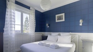 Camera da letto con un letto grande, biancheria da letto bianca, una finestra con tende leggere, un comodino con una lampada e un quadro appeso al muro.