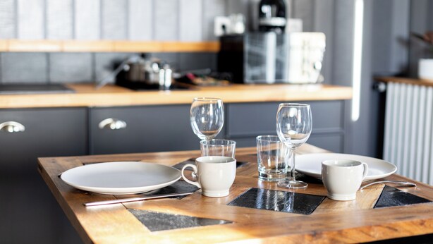 Un tavolo da pranzo apparecchiato con piatti, bicchieri e posate, con sullo sfondo una cucina completa di pensili ed elettrodomestici.