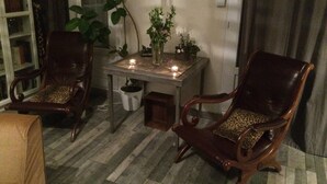 Quarto com duas cadeiras de madeira com almofadas de estampa de leopardo, uma mesinha com velas, um vaso de planta e uma estante de livros.