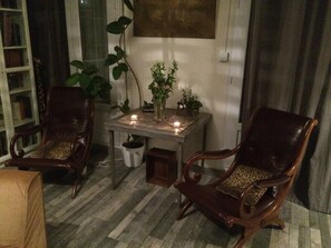 Ein Zimmer mit zwei Holzstühlen mit Leopardenmuster-Kissen, einem kleinen Tisch mit Kerzen, einer Topfpflanze und einem Bücherregal.