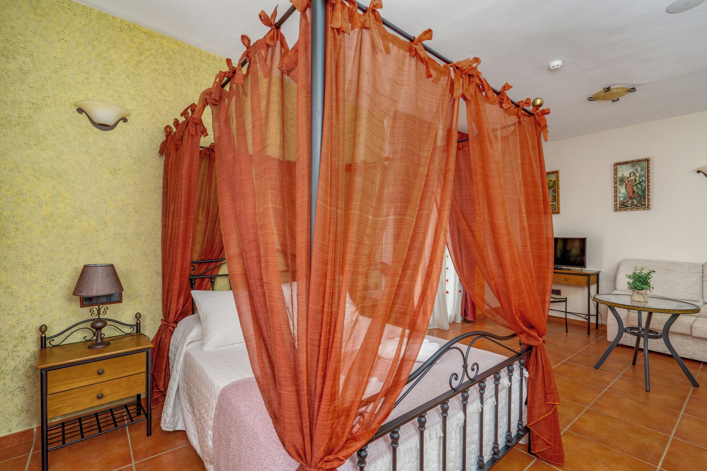 A bedroom with a four-poster bed, a bedside table with a lamp, a small table with a plant, and a sofa.