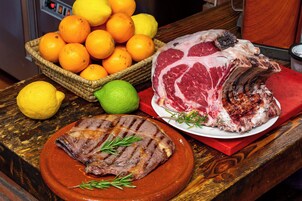 Una mesa con un plato de carne asada como pieza central, una bandeja de carne a la parrilla y una cesta de naranjas y limones.