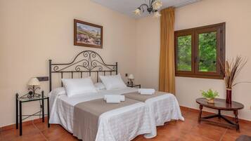 Cama doble con dos sábanas blancas, mesita de noche con lámpara, un cuadro en la pared y ventana con vistas a árboles.