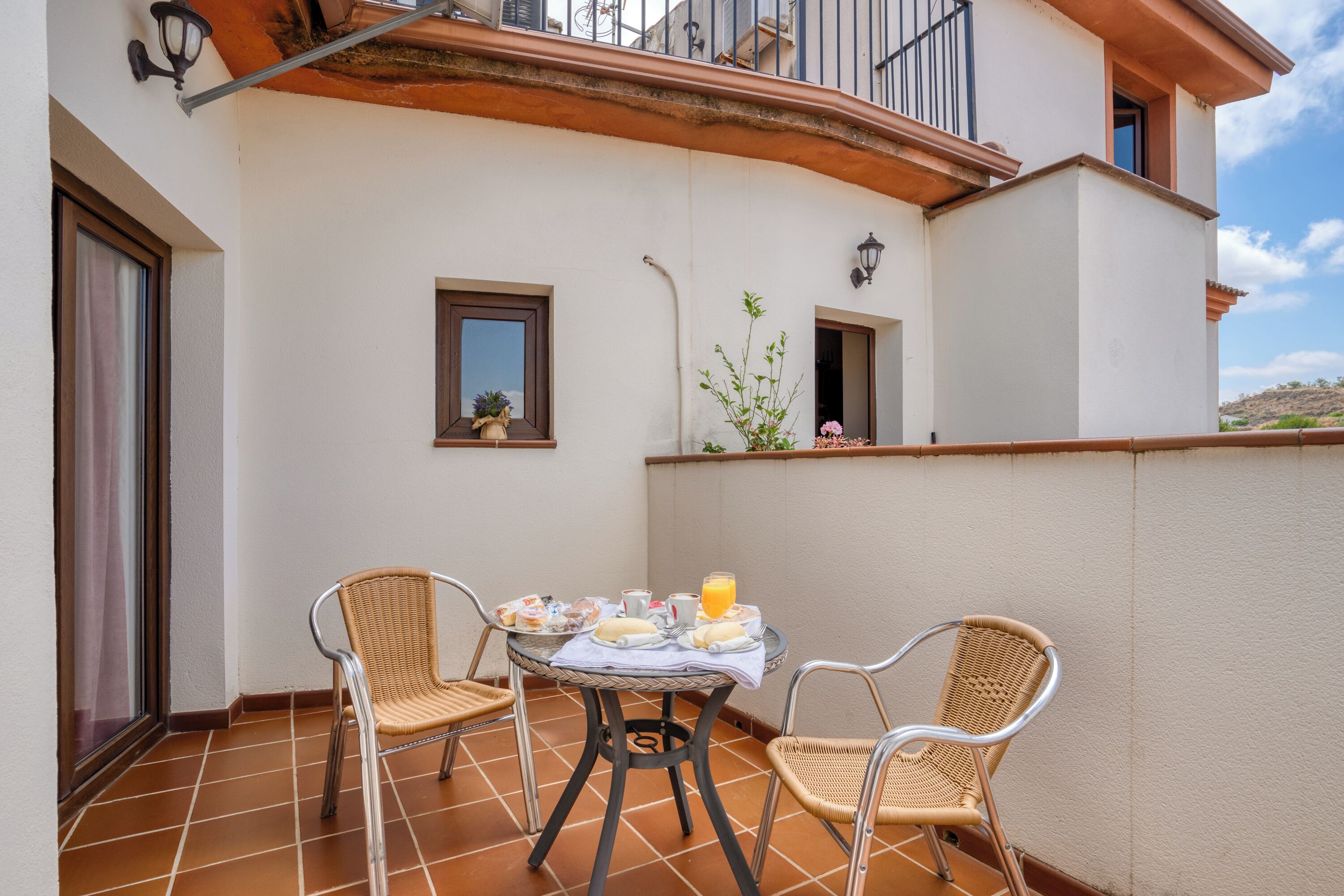 Una terraza con una mesa lista para el desayuno, dos sillas de mimbre y vistas del entorno.