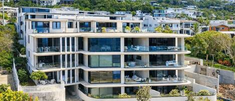 A multi-story residential building with balconies, a landscaped garden, and a hilly backdrop.