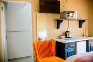 A hotel room with a bed, a TV mounted on the wall, a kitchenette with a coffee maker, and a chair.