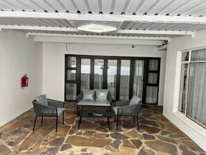 A covered patio with stone flooring, a sofa, two chairs, and a fire extinguisher.