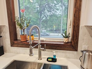 A kitchen with a window, a sink, a faucet, a potted plant, a soap dispenser, and a kettle.